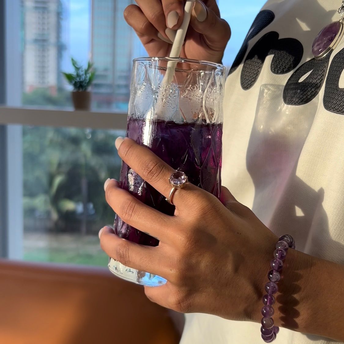 Person holding a glass of purple drink with a straw, wearing a white shirt with black designs and a bracelet.