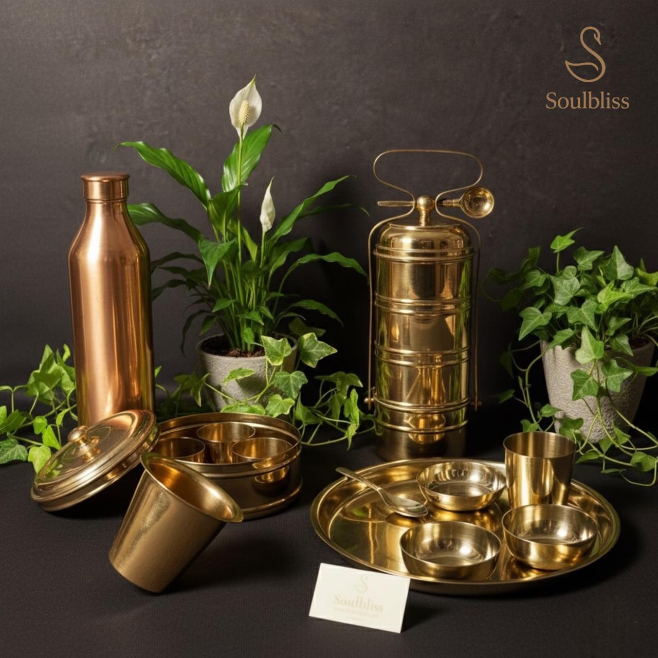 Traditional copper and peetal (brass) kitchen and dining set displayed on a black background, including a copper water bottle, brass tiffin lunchbox, brass thali plate, bowls, spoon, and glasses, styled with green indoor plants and Soulbliss branding.
