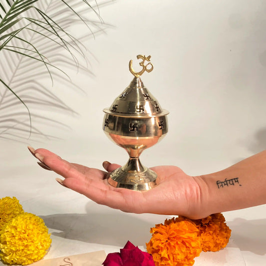 Hand holding a brass incense burner with flowers and a plant in the background