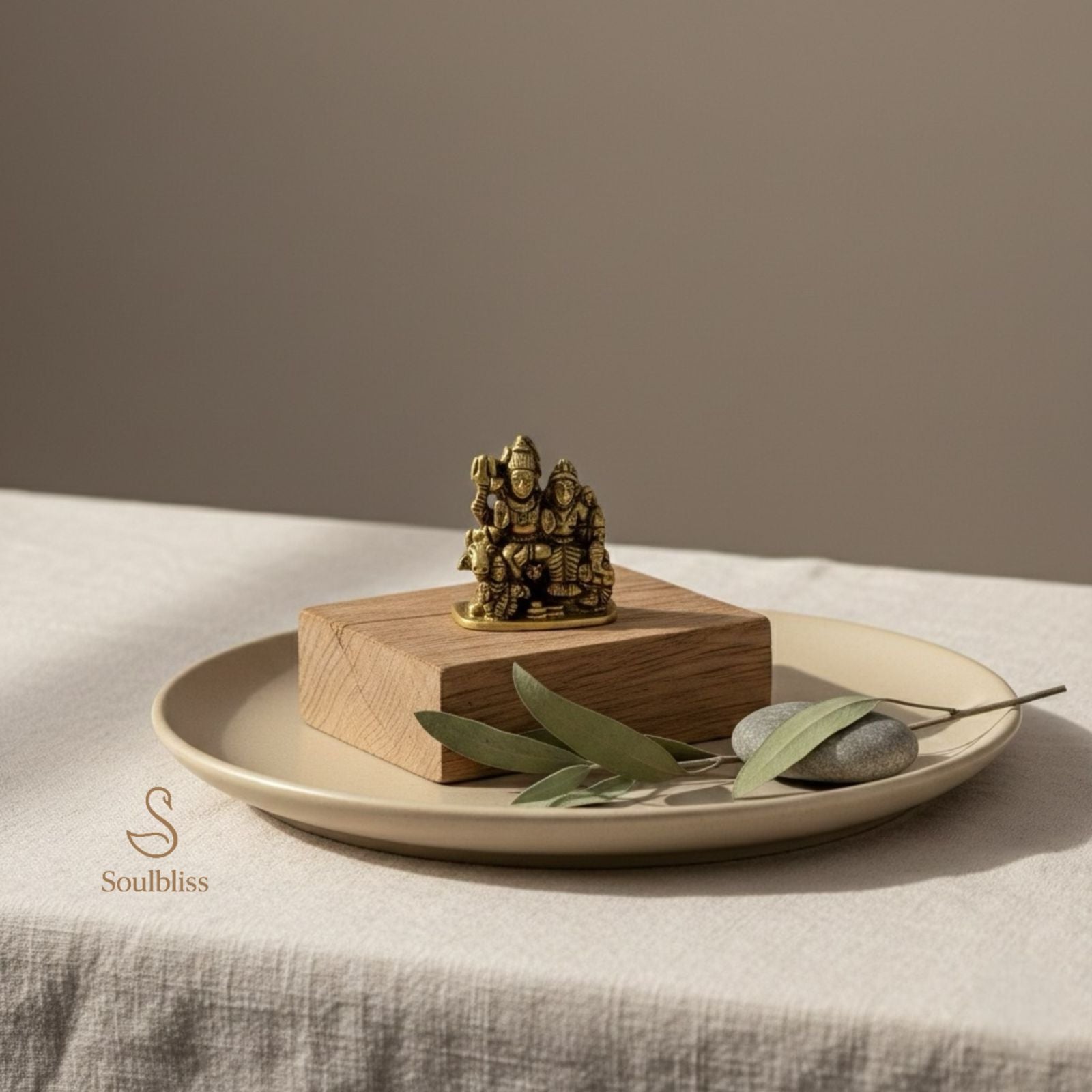 Small brass shiv parivar  statue on a wooden block with green leaves and stones on a beige plate against a neutral background.