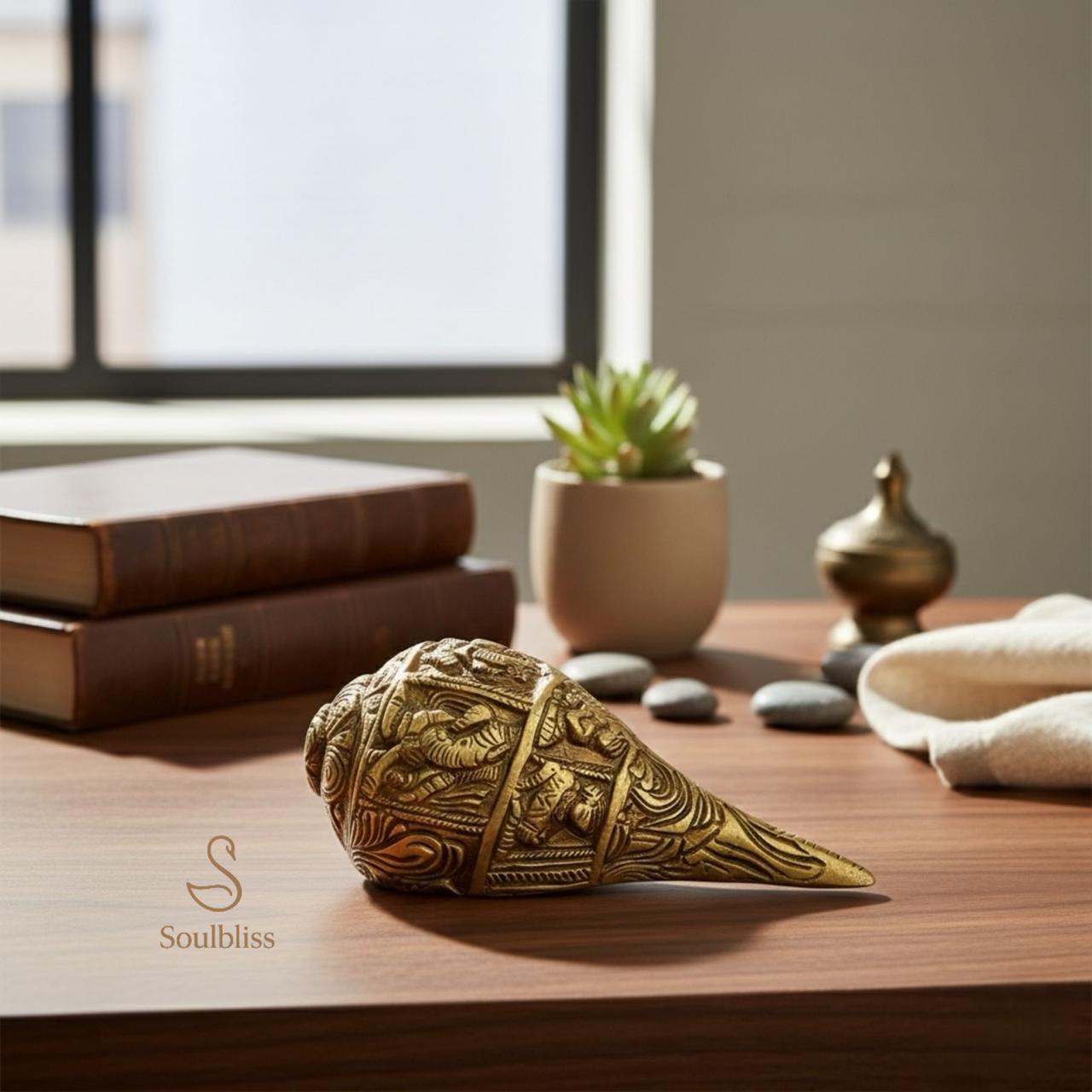 Decorative brass shankh used for vastu remedy on a wooden surface with books and a plant in the background.