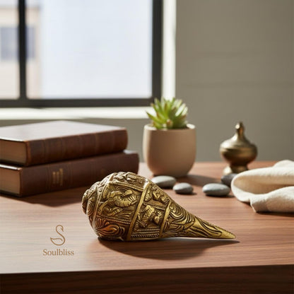 Decorative brass shankh used for vastu remedy on a wooden surface with books and a plant in the background.