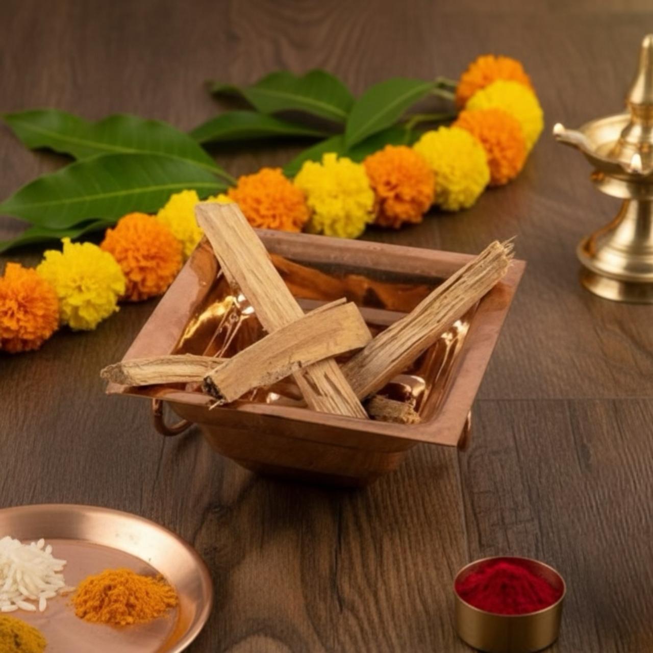 Incense sticks in a copper havan kund with marigold flowers, leaves, and a lamp on a wooden surface.