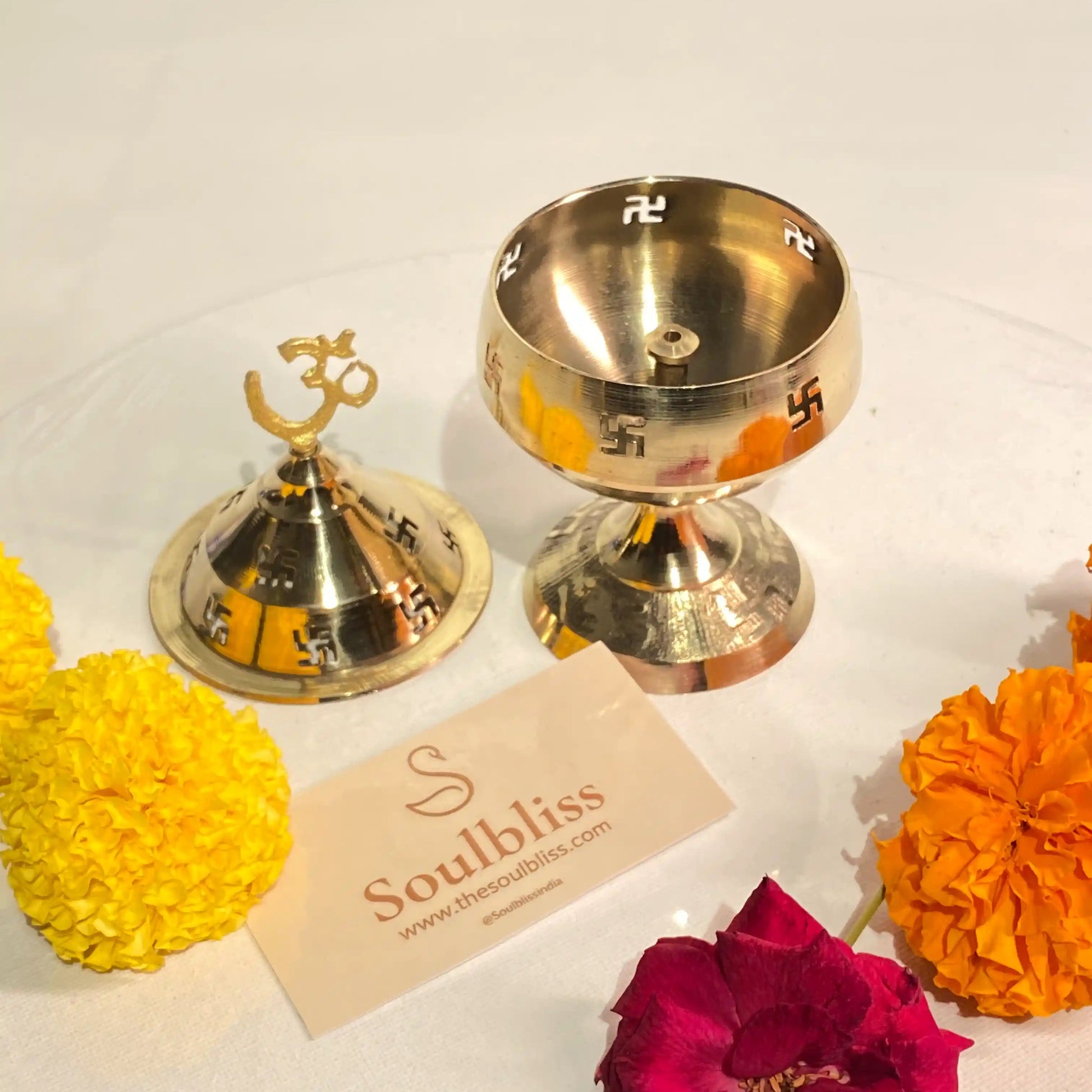 Two brass bells with a 'Soulbliss' card and colorful flowers on a white background