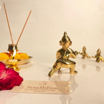 Gold figurine of a sitting laddu gopal  with incense sticks and flowers on a white surface, featuring the brand 'Soulbliss.'
